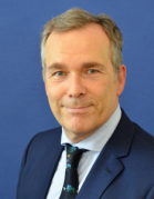 Prof. Dr. Heintz Mann mit grauem Haar trägt dunkelblauen Anzug, hellblaues Hemd und schwarze Krawatte mit buntem Muster vor blauem Hintergrund