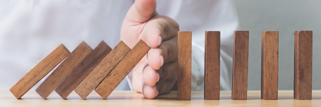 Hand stoppt fallende hölzerne Dominosteine, darunter das Wort 'PRÄVENTION' in orange.