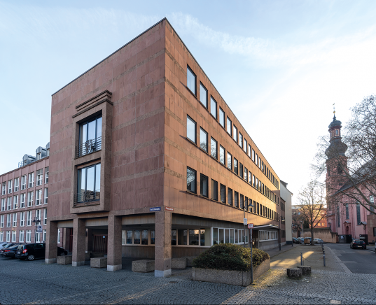 Der ehemalige Sitz der Landesärztekammer am Deutschhausplatz: ein eckiges, mehrstöckiges Gebäude mit brauner Fassade, im Hintergrund barocke Kirche mit Turm und Wetterfahne.